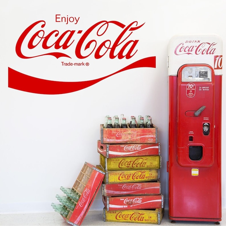 Puede incluir: Una m&aacute;quina expendedora de Coca-Cola vintage con un exterior rojo y un letrero blanco que dice "Drink Coca-Cola". La m&aacute;quina est&aacute; junto a una pila de cajas de madera llenas de botellas de vidrio de Coca-Cola. Las cajas est&aacute;n etiquetadas con el logotipo de Coca-Cola y las palabras "Drink Coca-Cola in Bottles".