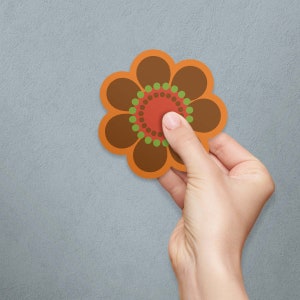 May include: A brown and orange flower-shaped sticker with a red, green, and brown center. The sticker is being placed on a gray wall.