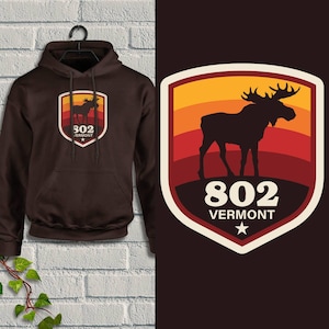May include: A brown hoodie with a front pocket and a drawstring hood. The hoodie features a shield-shaped graphic with a moose silhouette, the number 802, and the word "VERMONT". The graphic has a sunset color scheme.