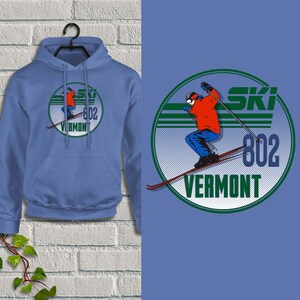 May include: A light blue hoodie with a graphic of a skier in red and blue, with the words "SKI 802 VERMONT" in a circular design. The hoodie is hanging on a black hanger against a white brick wall.