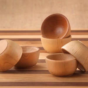 May include: A collection of small, wooden bowls. The bowls are light brown with a smooth finish, and some are stacked. They are arranged on a striped wooden surface, creating a natural and rustic aesthetic.