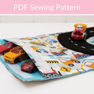 May include: A PDF sewing pattern for a toy car play mat. The mat features a road design and a pocket holding three toy cars. The fabric has a print of various vehicles. The text "PDF Sewing Pattern" is at the top.