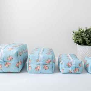 May include: A set of four rectangular zippered pouches in graduated sizes, featuring a light blue fabric base with a repeating pattern of pink roses and green leaves. Each pouch has a white zipper closure and a structured, boxy shape.