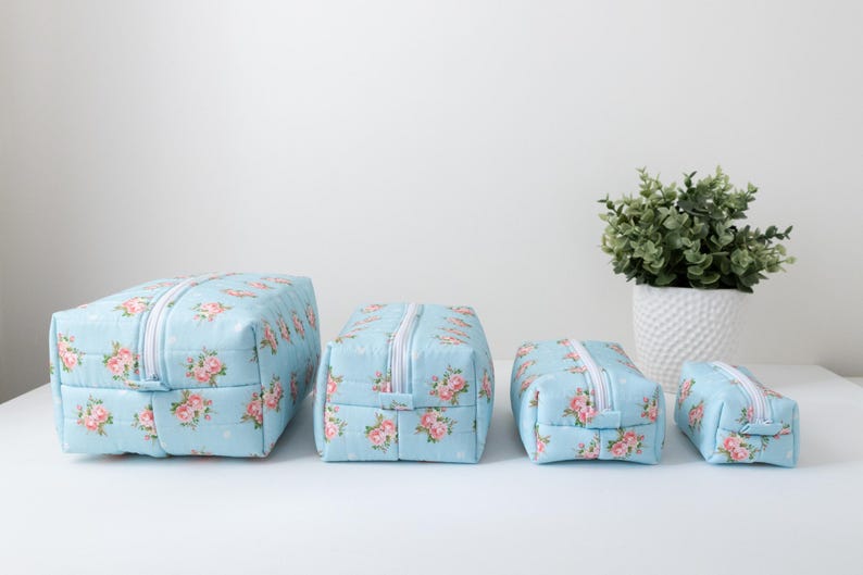 May include: A set of four rectangular zippered pouches in graduated sizes, featuring a light blue fabric base with a repeating pattern of pink roses and green leaves. Each pouch has a white zipper closure and a structured, boxy shape.