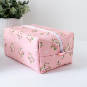 May include: A rectangular pink fabric pouch featuring a repeating pattern of small pink roses and white polka dots. The pouch has a white zipper closure along the top edge and a matching pink fabric zipper pull tab, showcasing a structured design.
