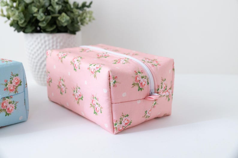 May include: A rectangular pink fabric pouch featuring a repeating pattern of small pink roses and white polka dots. The pouch has a white zipper closure along the top edge and a matching pink fabric zipper pull tab, showcasing a structured design.