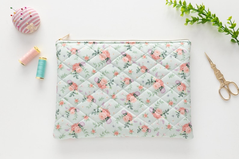 K&ouml;nnte beinhalten: Eine gesteppte Tasche mit floralem Muster aus rosa Rosen und gr&uuml;nen Bl&auml;ttern auf hellblauem Hintergrund, mit goldenem Rei&szlig;verschluss. N&auml;hutensilien, darunter ein Nadelkissen, Garnrollen und eine goldene Schere, sind um die Tasche herum angeordnet.
