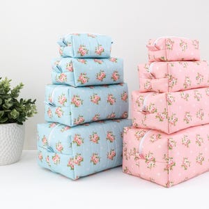 May include: A set of four light blue rectangular zippered pouches and four light pink rectangular zippered pouches, all featuring a white polka dot pattern and a floral print of small pink roses with green leaves, stacked in two separate columns.