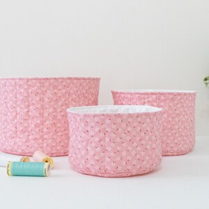 May include: Three pink fabric storage baskets of varying sizes, with a floral pattern and white lining. Several spools of thread in various colors are in front of the baskets. The baskets are on a white surface.