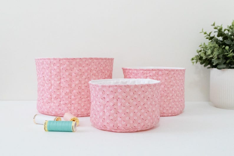 May include: Three pink fabric storage baskets of varying sizes, with a floral pattern and white lining. Several spools of thread in various colors are in front of the baskets. The baskets are on a white surface.