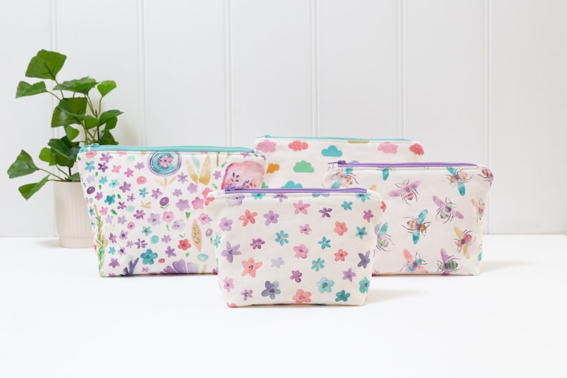 May include: Four zippered pouches with different floral and bee patterns. The pouches are made of white fabric with a light blue zipper and a purple zipper pull.