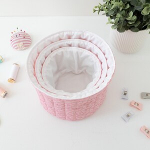 May include: A set of three pink fabric nesting baskets with white lining. The image also shows sewing supplies, including spools of thread, a pincushion, gold scissors, and fabric clips. A potted plant is in the background.
