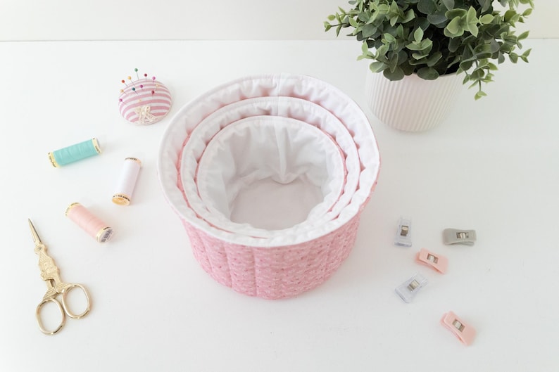 May include: A set of three pink fabric nesting baskets with white lining. The image also shows sewing supplies, including spools of thread, a pincushion, gold scissors, and fabric clips. A potted plant is in the background.