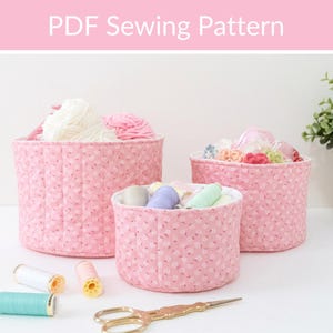May include: Three pink fabric storage baskets of varying sizes, filled with yarn, thread, and sewing notions. The baskets have a floral pattern and are lined with white fabric. The image includes spools of thread and a pair of gold scissors. Text at the top reads "PDF Sewing Pattern."