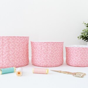 May include: Three pink fabric storage baskets of varying sizes, with a floral pattern. Sewing supplies, including spools of thread and gold scissors, are arranged in front of the baskets. A small potted plant is in the background.