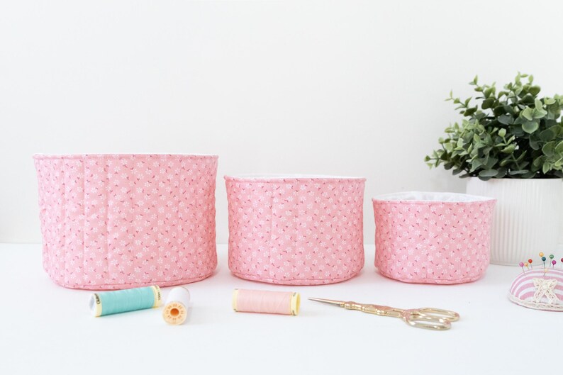 May include: Three pink fabric storage baskets of varying sizes, with a floral pattern. Sewing supplies, including spools of thread and gold scissors, are arranged in front of the baskets. A small potted plant is in the background.