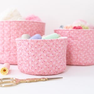 May include: Three pink fabric storage baskets with white floral patterns, filled with yarn and thread. Gold scissors and spools of thread are in the foreground. The baskets are different sizes, perfect for organizing craft supplies.