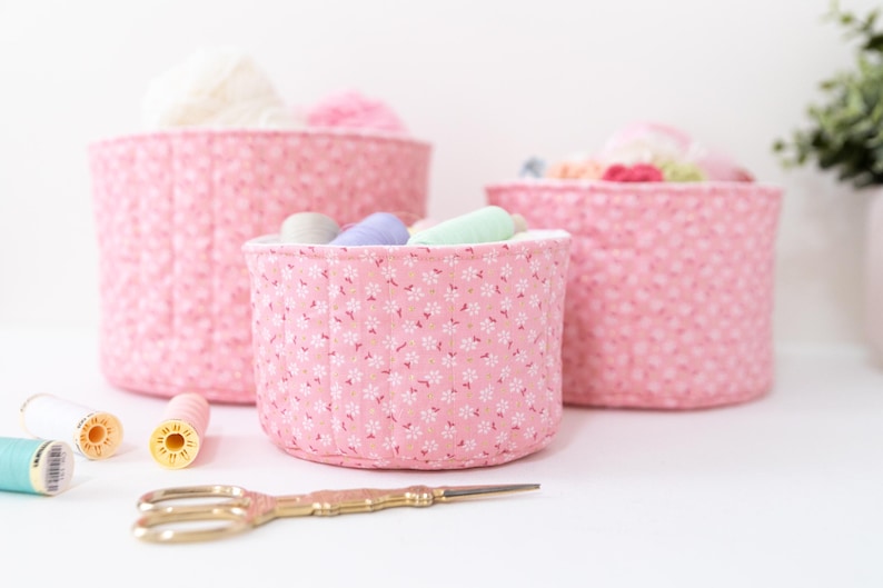 May include: Three pink fabric storage baskets with white floral patterns, filled with yarn and thread. Gold scissors and spools of thread are in the foreground. The baskets are different sizes, perfect for organizing craft supplies.