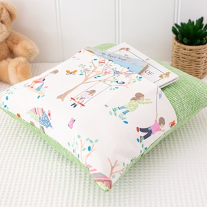 Puede incluir: Una almohada verde y blanca con un bolsillo para un libro. La almohada tiene un estampado de ni&ntilde;os jugando en un parque. El libro en el bolsillo se titula "The Book of"