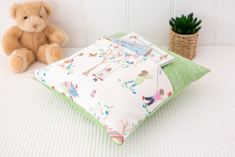 Puede incluir: Una almohada verde y blanca con un bolsillo para un libro. La almohada tiene un estampado de ni&ntilde;os jugando en un parque. El libro en el bolsillo se titula "The Book of"