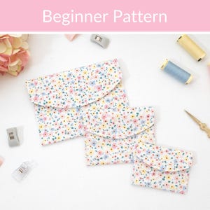 May include: Three fabric pouches in varying sizes featuring a white background with a small floral pattern in pink, yellow, and blue. The pouches have rounded envelope-style flaps. Sewing supplies including thread spools and scissors are visible.