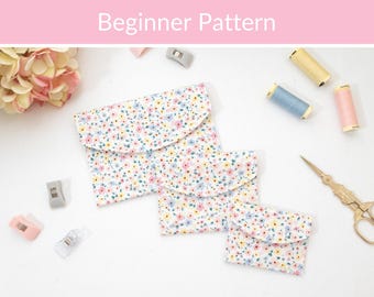May include: Three fabric pouches in varying sizes featuring a white background with a small floral pattern in pink, yellow, and blue. The pouches have rounded envelope-style flaps. Sewing supplies including thread spools and scissors are visible.