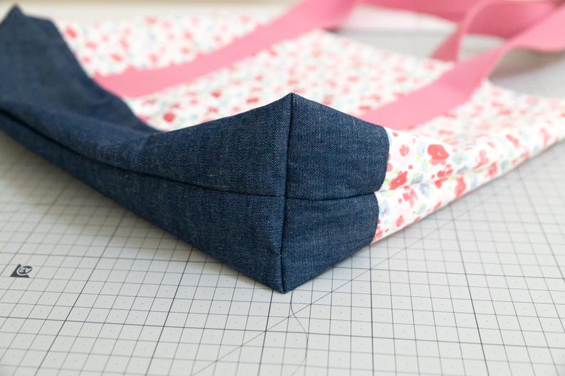 Puede incluir: Un bolso tote que combina tela de mezclilla azul oscuro en la base y los laterales con un panel de tela blanca estampado con un peque&ntilde;o patr&oacute;n floral en rojo y azul claro. El bolso incluye dos correas largas de tela en color rosa s&oacute;lido.