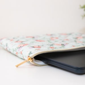 K&ouml;nnte beinhalten: Eine Laptop-H&uuml;lle mit floralem Muster und goldenem Rei&szlig;verschluss, teilweise ge&ouml;ffnet, um einen schwarzen Laptop zu zeigen. Die H&uuml;lle hat einen hellblauen Hintergrund mit rosa und lila Blumendesigns.