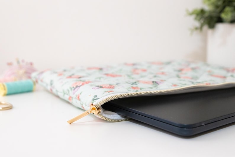 K&ouml;nnte beinhalten: Eine Laptop-H&uuml;lle mit floralem Muster und goldenem Rei&szlig;verschluss, teilweise ge&ouml;ffnet, um einen schwarzen Laptop zu zeigen. Die H&uuml;lle hat einen hellblauen Hintergrund mit rosa und lila Blumendesigns.