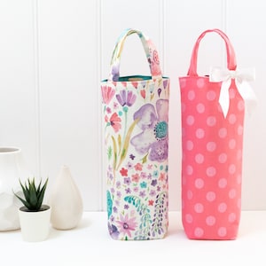 May include: Two wine bottle bags. One is a floral print with purple, pink, and blue flowers. The other is pink with white polka dots and a white bow. Both have handles.