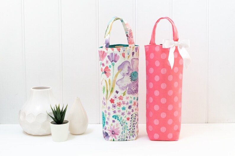 May include: Two wine bottle bags. One is a floral print with purple, pink, and blue flowers. The other is pink with white polka dots and a white bow. Both have handles.