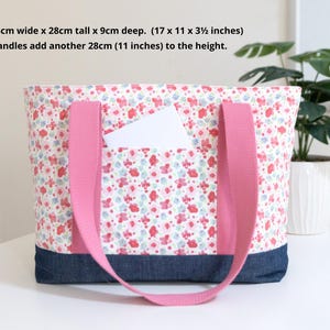 Puede incluir: Un bolso tote con estampado floral en rosa, rojo y azul, con una base de mezclilla azul oscuro y asas de tela rosa. Mide 43 cm de ancho por 28 cm de alto y 9 cm de profundidad, con asas que a&ntilde;aden otros 28 cm a la altura total.