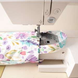 May include: A white sewing machine stitching a floral patterned fabric. The fabric features purple, pink, and blue watercolor flowers on a white background. The machine's needle is in action, creating a seam.