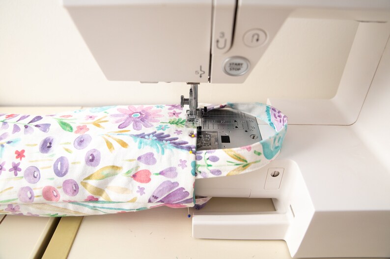 May include: A white sewing machine stitching a floral patterned fabric. The fabric features purple, pink, and blue watercolor flowers on a white background. The machine's needle is in action, creating a seam.