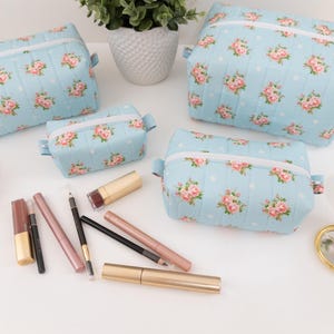 May include: Three light blue quilted cosmetic bags featuring a pink rose and white polka dot pattern with white zippers. The set includes various makeup items such as lipsticks, eyeliners, and a small gold compact mirror arranged on a surface.
