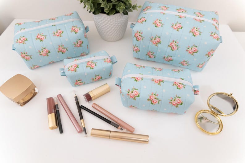 May include: Three light blue quilted cosmetic bags featuring a pink rose and white polka dot pattern with white zippers. The set includes various makeup items such as lipsticks, eyeliners, and a small gold compact mirror arranged on a surface.