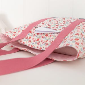 Puede incluir: Un bolso tote con un estampado floral de flores rojas, azules y verdes sobre fondo blanco. El bolso tiene dos asas largas de tela rosa y un panel lateral azul oscuro. Una peque&ntilde;a tarjeta rectangular blanca est&aacute; metida en un bolsillo exterior.