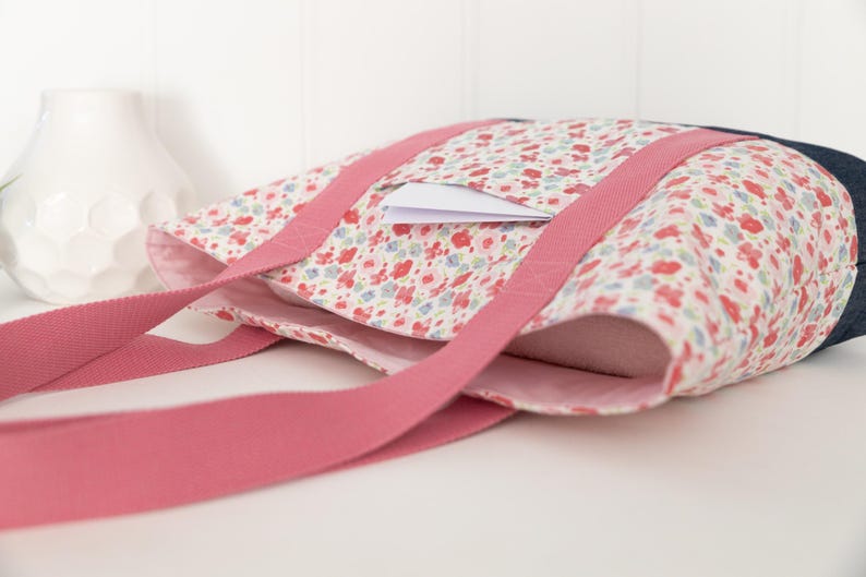 Puede incluir: Un bolso tote con un estampado floral de flores rojas, azules y verdes sobre fondo blanco. El bolso tiene dos asas largas de tela rosa y un panel lateral azul oscuro. Una peque&ntilde;a tarjeta rectangular blanca est&aacute; metida en un bolsillo exterior.