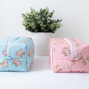 May include: Two rectangular zippered pouches, one in light blue and one in light pink, both featuring a repeating floral pattern of small pink roses and green leaves with a white zipper closure running along the top center of each bag.