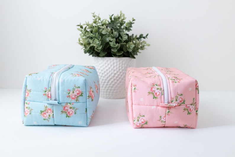 May include: Two rectangular zippered pouches, one in light blue and one in light pink, both featuring a repeating floral pattern of small pink roses and green leaves with a white zipper closure running along the top center of each bag.
