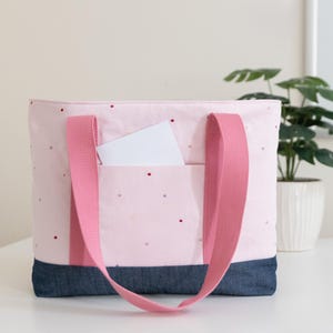 Puede incluir: Un bolso tote de color rosa claro con un patr&oacute;n de peque&ntilde;os puntos rojos y dorados, una base de mezclilla azul oscuro y dos asas anchas de tela rosa. Una tarjeta rectangular blanca est&aacute; metida en el bolsillo exterior delantero.
