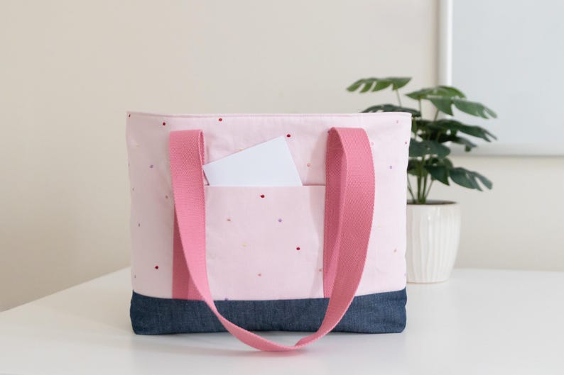 Puede incluir: Un bolso tote de color rosa claro con un patr&oacute;n de peque&ntilde;os puntos rojos y dorados, una base de mezclilla azul oscuro y dos asas anchas de tela rosa. Una tarjeta rectangular blanca est&aacute; metida en el bolsillo exterior delantero.