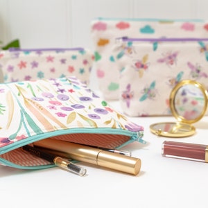 May include: A set of four floral patterned zippered pouches with a variety of designs, including flowers, bees, and clouds. The pouches are made of a soft, white fabric and have a light purple zipper. The pouches are arranged on a white surface with a small gold mirror and a tube of lipstick.