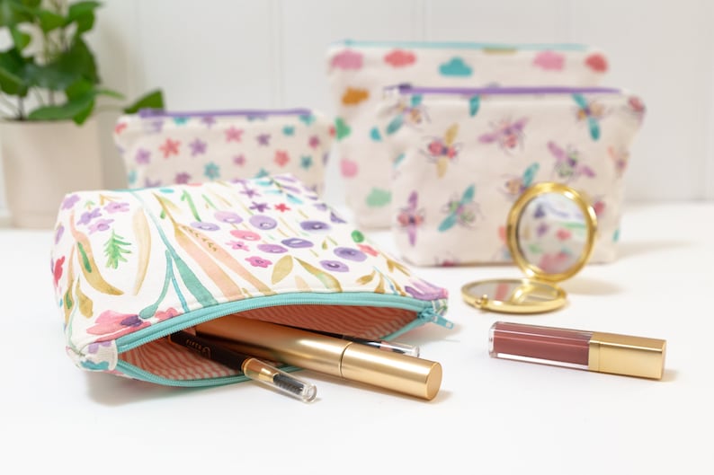 May include: A set of four floral patterned zippered pouches with a variety of designs, including flowers, bees, and clouds. The pouches are made of a soft, white fabric and have a light purple zipper. The pouches are arranged on a white surface with a small gold mirror and a tube of lipstick.