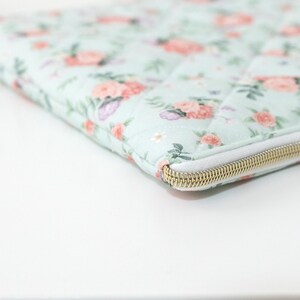 K&ouml;nnte beinhalten: Eine gesteppte Laptoptasche mit einem Blumenmuster in Rosa, Lila und Gr&uuml;n auf hellblauem Hintergrund. Die Tasche hat einen wei&szlig;en Rei&szlig;verschluss mit einem goldfarbenen Metallschieber.