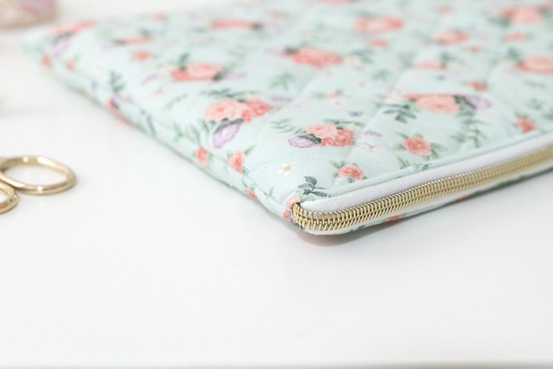 K&ouml;nnte beinhalten: Eine gesteppte Laptoptasche mit einem Blumenmuster in Rosa, Lila und Gr&uuml;n auf hellblauem Hintergrund. Die Tasche hat einen wei&szlig;en Rei&szlig;verschluss mit einem goldfarbenen Metallschieber.
