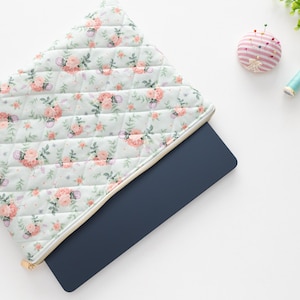 K&ouml;nnte beinhalten: Eine gesteppte Laptoptasche mit einem floralen Muster in Rosa- und Pfirsicht&ouml;nen und einem goldenen Rei&szlig;verschluss. Ein marineblauer Laptop ist teilweise sichtbar. N&auml;hutensilien, darunter ein Nadelkissen, Garnrollen und eine Schere, sind in der N&auml;he angeordnet.