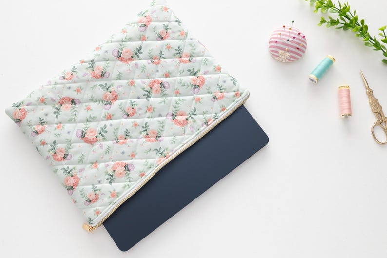 K&ouml;nnte beinhalten: Eine gesteppte Laptoptasche mit einem floralen Muster in Rosa- und Pfirsicht&ouml;nen und einem goldenen Rei&szlig;verschluss. Ein marineblauer Laptop ist teilweise sichtbar. N&auml;hutensilien, darunter ein Nadelkissen, Garnrollen und eine Schere, sind in der N&auml;he angeordnet.