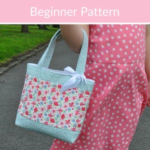 May include: A light blue tote bag with a white polka dot pattern and a central horizontal panel featuring a floral print of pink, red, and blue flowers. The bag includes two matching handles and a white ribbon bow tied to one side. Text reads Beginner Pattern.