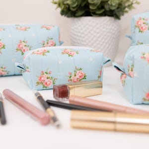 May include: Three light blue quilted cosmetic bags featuring a repeating pattern of pink roses and white polka dots, each with a white zipper closure. Several makeup items, including lipsticks and eyeliner pencils, are arranged in the foreground.